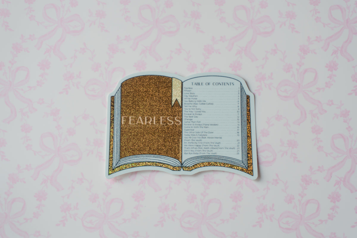 A die-cut sticker shaped like an open book with a gold glittery cover and a gold-accented page, placed on a light pink patterned background. The sticker has a faint 'FEARLESS' watermark across the center.