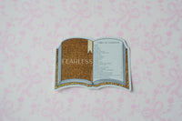 A die-cut sticker shaped like an open book with a gold glittery cover and a gold-accented page, placed on a light pink patterned background. The sticker has a faint 'FEARLESS' watermark across the center.