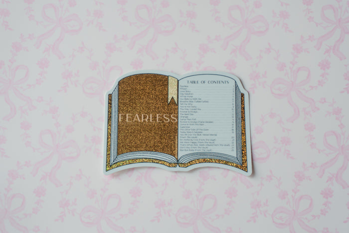A die-cut sticker shaped like an open book with a gold glittery cover and a gold-accented page, placed on a light pink patterned background. The sticker has a faint 'FEARLESS' watermark across the center.
