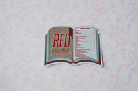 A die-cut sticker shaped like an open book titled 'RED TAYLOR'S VERSION' with a glittery gold interior page and a table of contents on the right page, shown on a light pink patterned background.