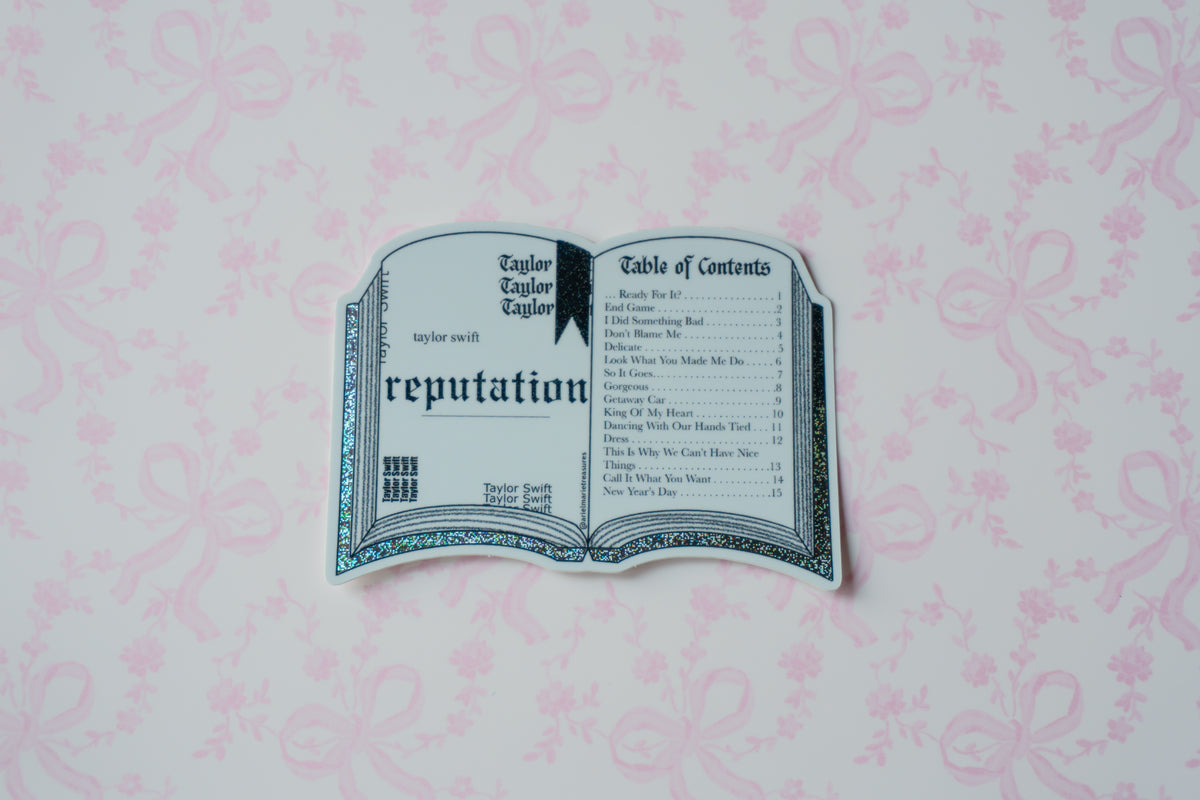 A die-cut sticker shaped like an open book titled 'reputation' from Taylor Swift's 'Fearless (Taylor's Version)' album, displayed on a light pink patterned background.