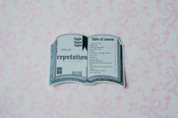 A die-cut sticker shaped like an open book titled 'reputation' from Taylor Swift's 'Fearless (Taylor's Version)' album, displayed on a light pink patterned background.