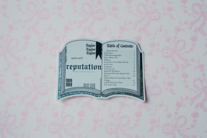 A die-cut sticker shaped like an open book titled 'reputation' from Taylor Swift's 'Fearless (Taylor's Version)' album, displayed on a light pink patterned background.