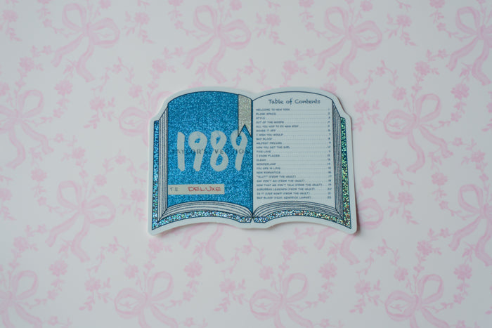 A die-cut sticker shaped like an open book, featuring a glittery blue cover with '1989' in white and a table of contents page, placed on a light pink patterned background.