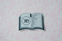 A novelty book-shaped sticker or decal featuring 'The Tortured Poets Department' logo and a table of contents design, placed on a light pink patterned background.