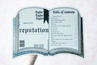 A novelty sticker or decal shaped like an open book, featuring Taylor Swift's 'Reputation' album cover art and printed table of contents text, held against a pale pink background.
