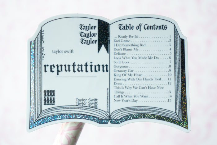 A novelty sticker or decal shaped like an open book, featuring Taylor Swift's 'Reputation' album cover art and printed table of contents text, held against a pale pink background.