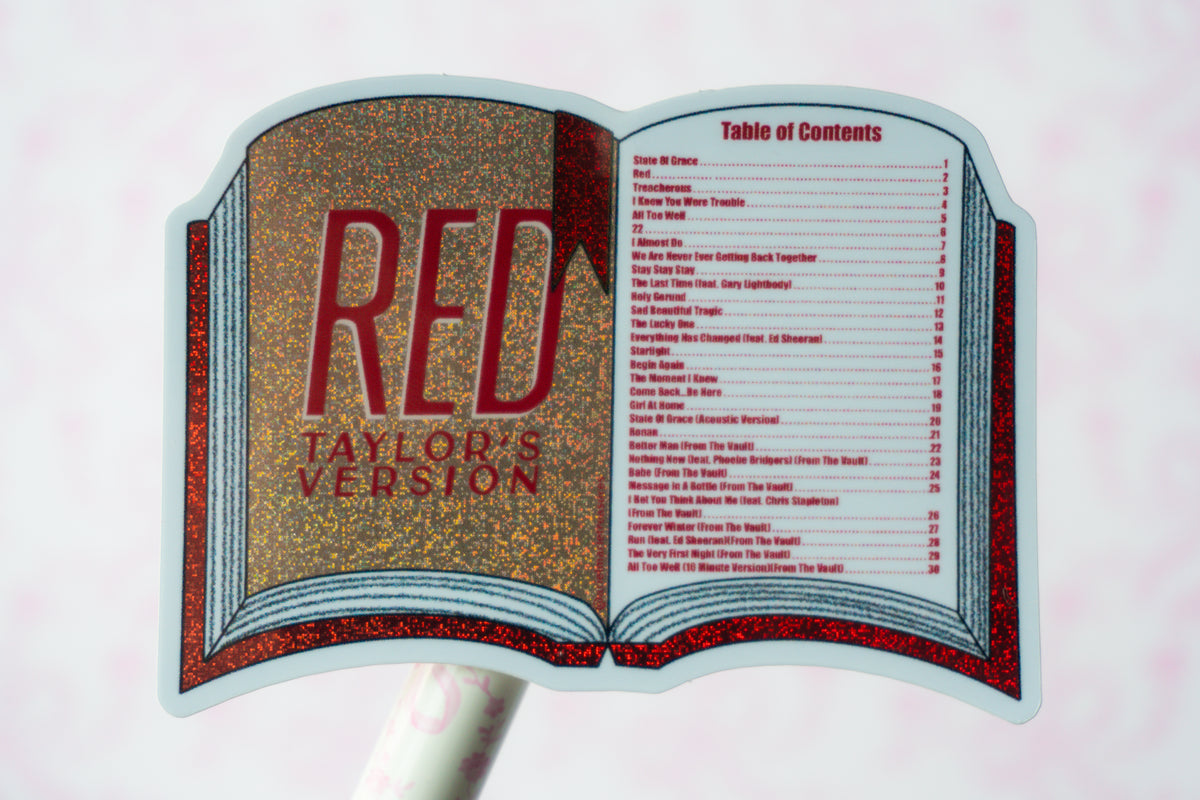 A die-cut sticker or sign shaped like an open book, featuring glittery gold and red artwork with the text 'RED TAYLOR'S VERSION' and a printed table of contents on the right page, shown against a pale pink background.