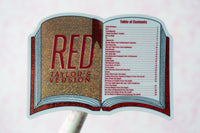 A die-cut sticker or sign shaped like an open book, featuring glittery gold and red artwork with the text 'RED TAYLOR'S VERSION' and a printed table of contents on the right page, shown against a pale pink background.