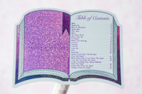 A glittery open book-shaped candle with a purple glittery left page, a dark purple spine, and a light blue right page printed with a table of contents, shown on a white background.