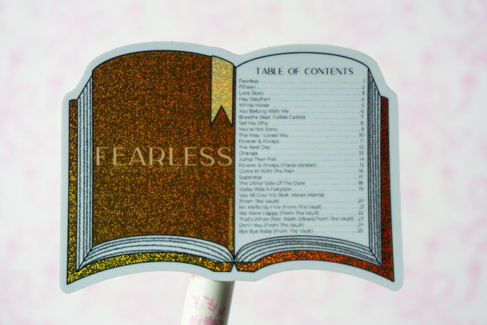 A novelty book-shaped sign featuring an open book design with glittery brown and gold textures, a printed table of contents, and the word 'FEARLESS' on the left page, mounted on a white stick against a pale pink background.