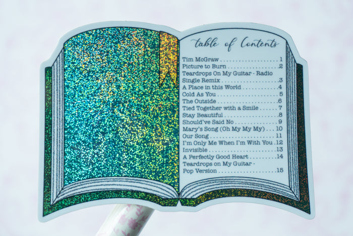 A glittery open book-shaped sticker or decal featuring a teal and gold sparkling cover and printed song-list style text on the pages, shown against a pale background.