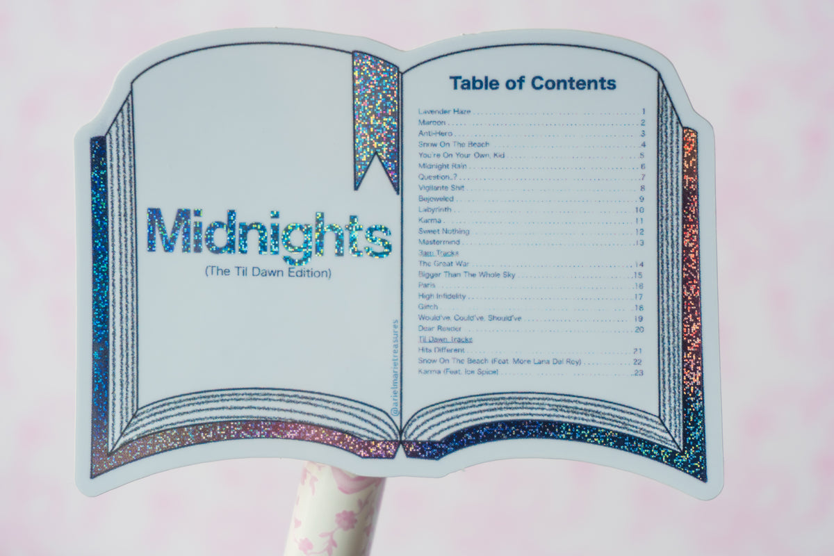 A book-shaped sign or stand featuring an open book design with the title 'Midnights (The Ti Dawn Edition)' and a printed table of contents, mounted on a pink-and-white striped pole against a soft pink background.