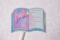 A book-shaped table topper or sticker with a glittery pink and blue cover design and a printed table of contents on the pages, mounted on a white stick against a pink patterned background.