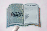 A die-cut sticker shaped like an open book titled 'folklore (deluxe version)' with a glittery blue-gray cover and an orange-gold patterned spine, shown on a white stick against a pale pink background.
