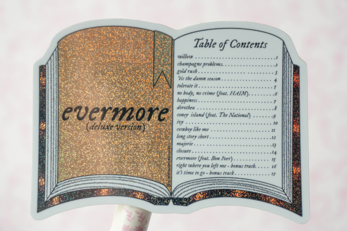 A book-shaped sticker or decal featuring an open book design with the title 'evermore ( deluxe version)' on the left page and a 'Table of Contents' on the right page, shown against a pale pink background.