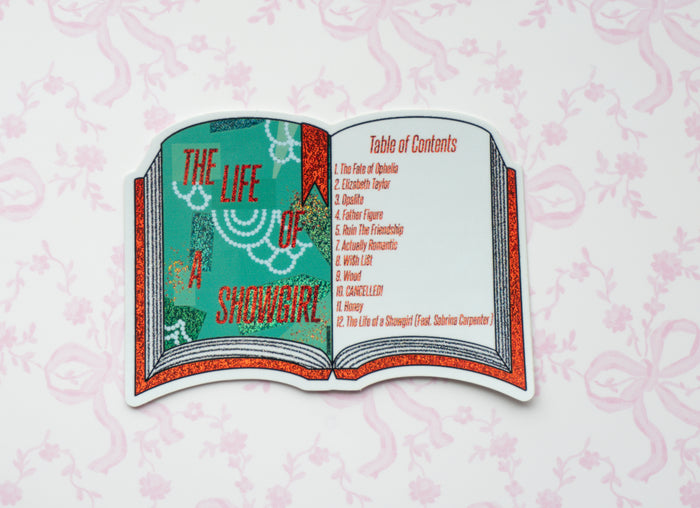 A die-cut sticker shaped like an open book featuring the title 'The Life of a Showgirl' on the left page and a 'Table of Contents' on the right page, styled with a glittery red and teal design, displayed on a light pink patterned background.