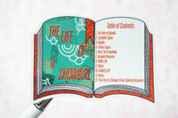 A close-up of an open book-shaped sticker or decal showing the cover art and table of contents for 'The Life of a Showgirl' on a pale pink background.