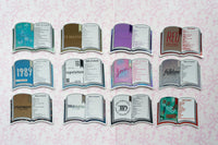 A set of 12 book-shaped stickers arranged in three rows on a light pink patterned background. The stickers depict various classic and popular books with colorful covers, including '1989', 'Reputation', 'lover', 'folklore', 'Evermore', 'Midnights', 'The Third Floor', 'Midnight', 'The Power', 'The Road', and 'Red'.