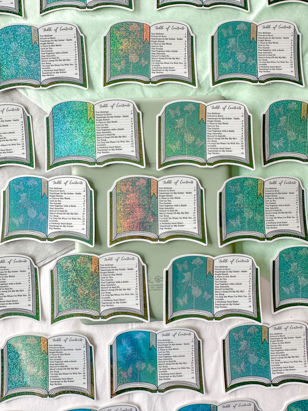 A repeating pattern of book-shaped stickers arranged on a pale mint background. Each card has a teal and multicolor glittery cover with an open-page interior showing the text 'Table of Contents' and a list of names.