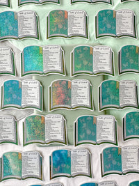 A repeating pattern of book-shaped stickers arranged on a pale mint background. Each card has a teal and multicolor glittery cover with an open-page interior showing the text 'Table of Contents' and a list of names.