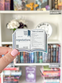 A hand with pink nail polish holds a miniature book-shaped sticker or card featuring Taylor Swift's 'reputation' table of contents, with a blurred bookshelf in the background.