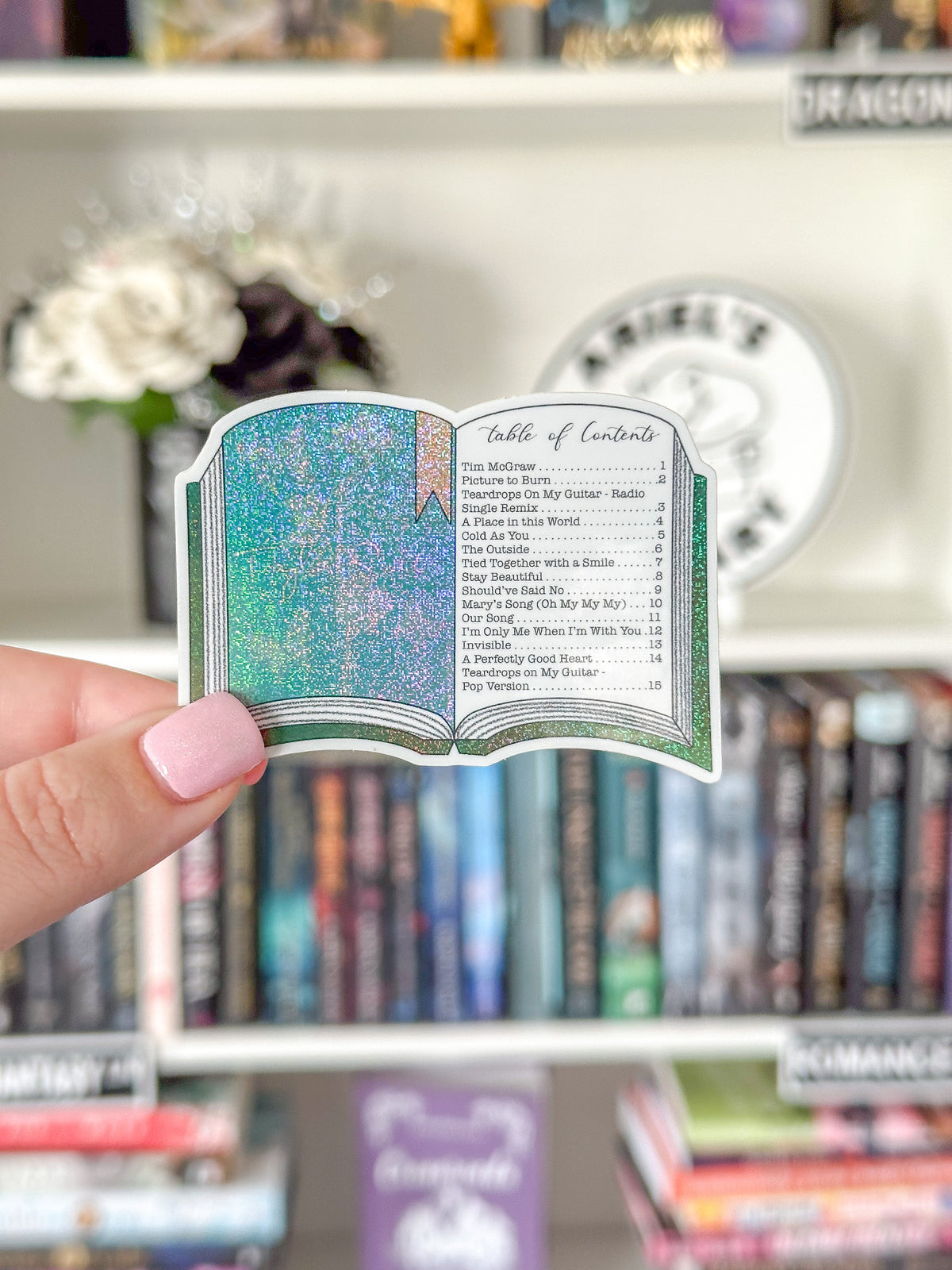 A hand with pink nail polish holds a die-cut sticker shaped like an open book showing a glittery teal and blue table of contents, with a blurred bookshelf background.