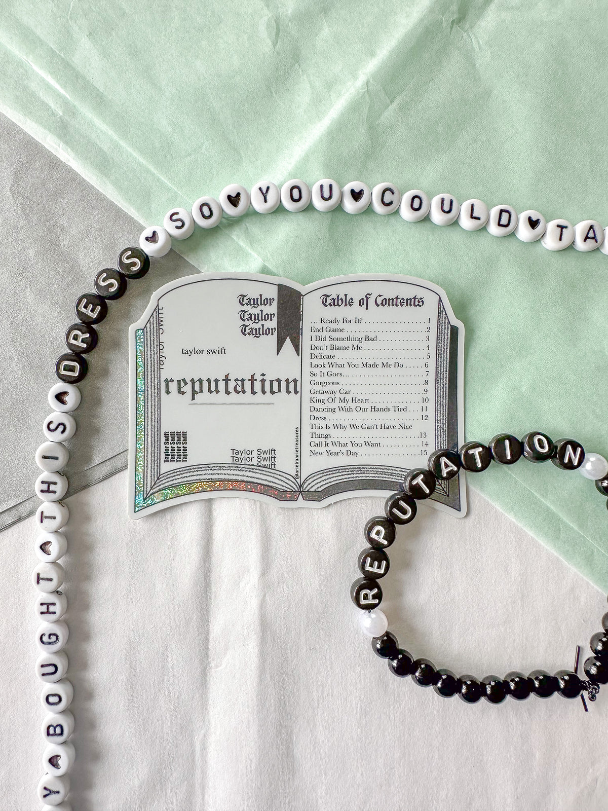 A beaded necklace and matching beaded bracelet styled on a white and mint green fabric background, featuring black and white beads spelling out Taylor Swift's 'Reputation' album artwork and track names.