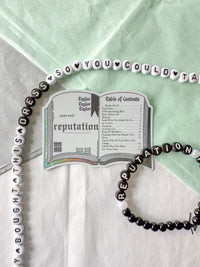 A beaded necklace and matching beaded bracelet styled on a white and mint green fabric background, featuring black and white beads spelling out Taylor Swift's 'Reputation' album artwork and track names.