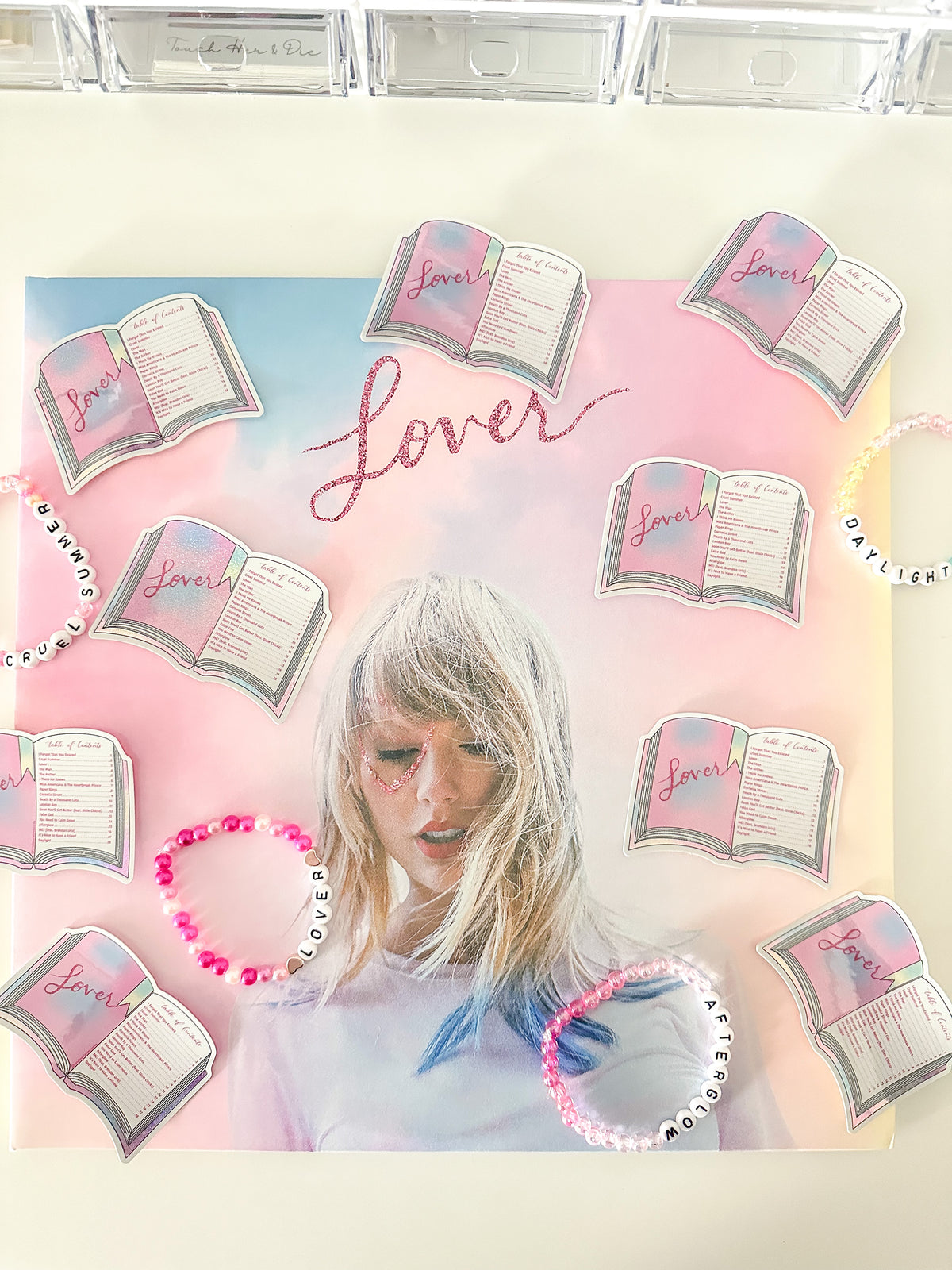 A square canvas print featuring a pastel pink and blue gradient background with the word 'Lover' in cursive, surrounded by illustrated open books and a portrait of Taylor Swift. Several beaded bracelets with matching book and Taylor Swift designs are arranged around the canvas on a light surface.