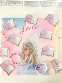 A square canvas print featuring a pastel pink and blue gradient background with the word 'Lover' in cursive, surrounded by illustrated open books and a portrait of Taylor Swift. Several beaded bracelets with matching book and Taylor Swift designs are arranged around the canvas on a light surface.