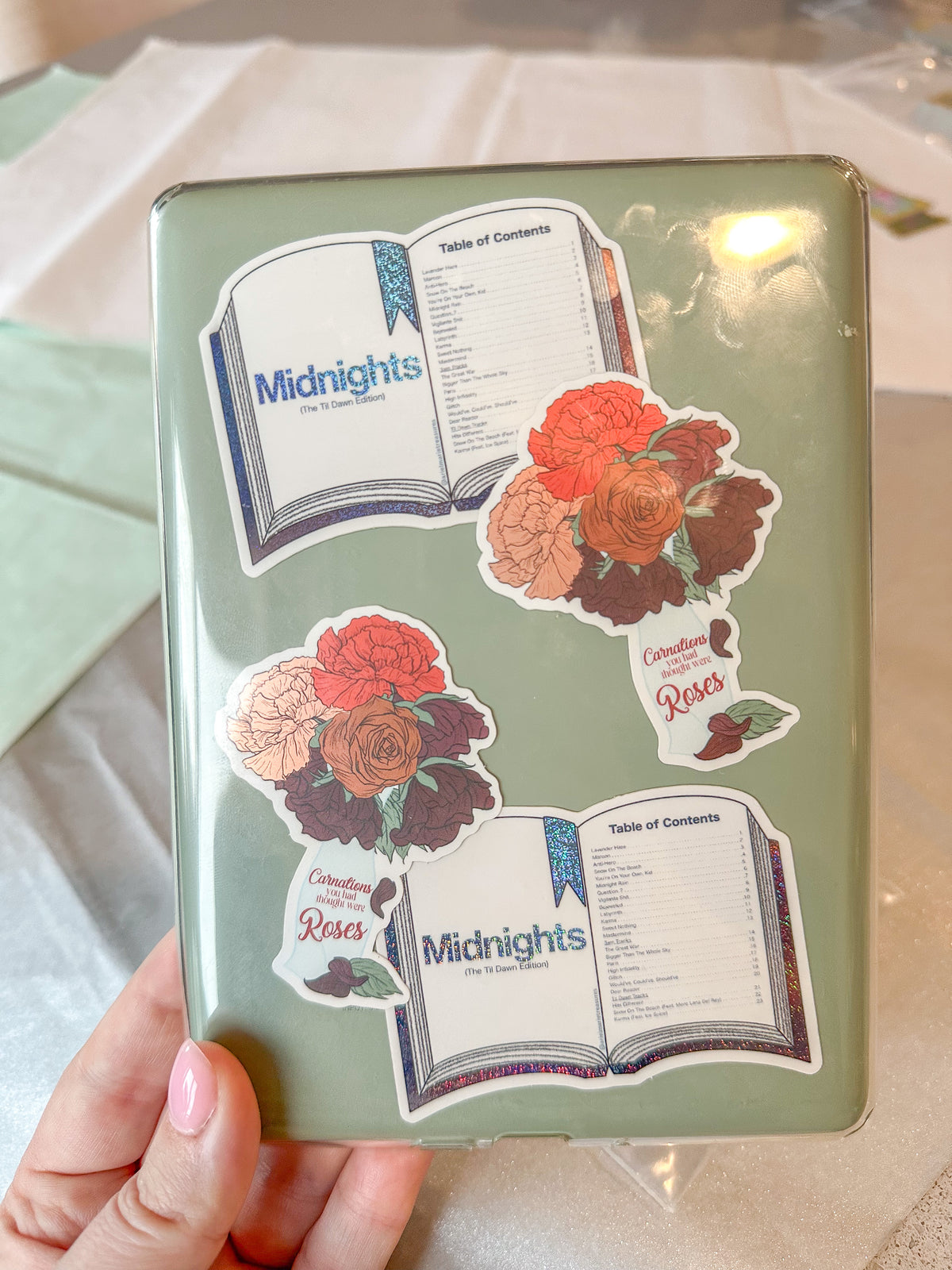 A hand holds a mint green hardcover notebook decorated with four decorative stickers featuring roses and open books with the title 'Midnights'.