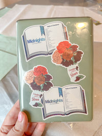 A hand holds a mint green hardcover notebook decorated with four decorative stickers featuring roses and open books with the title 'Midnights'.