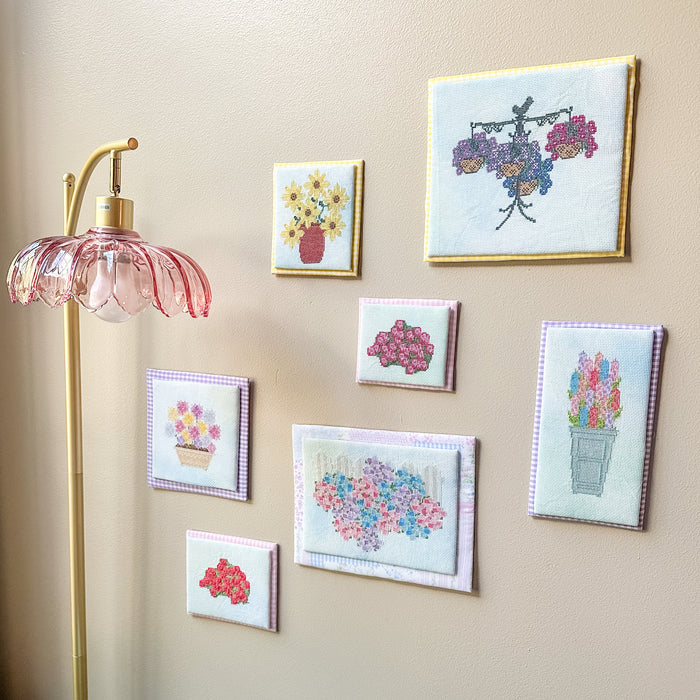 A gallery wall display of seven small framed floral needlepoint or embroidery art pieces in various colors, mounted on a beige wall next to a gold floor lamp with a pink glass shade.