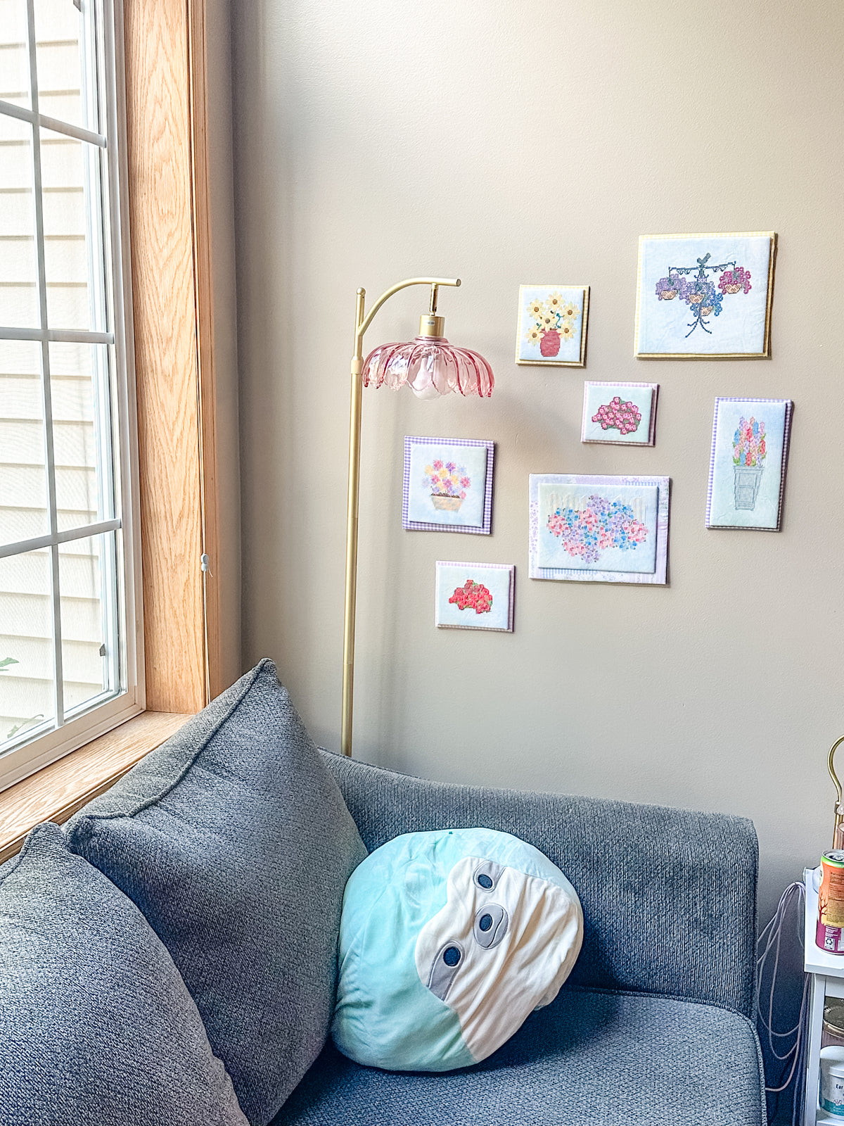 A cozy living room corner with a gray fabric sofa, a round light blue and cream plush pillow, a gold floor lamp with a pink bell-shaped shade, and a gallery wall of small framed floral art above.