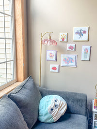 A cozy living room corner with a gray fabric sofa, a round light blue and cream plush pillow, a gold floor lamp with a pink bell-shaped shade, and a gallery wall of small framed floral art above.