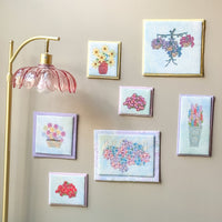 A gallery wall display of seven small framed floral art prints in various colors and styles, arranged on a beige wall next to a gold floor lamp.