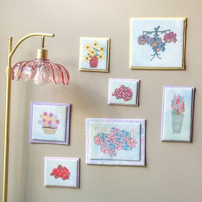 A gallery wall display of seven small framed floral art prints in various colors and styles, arranged on a beige wall next to a gold floor lamp.