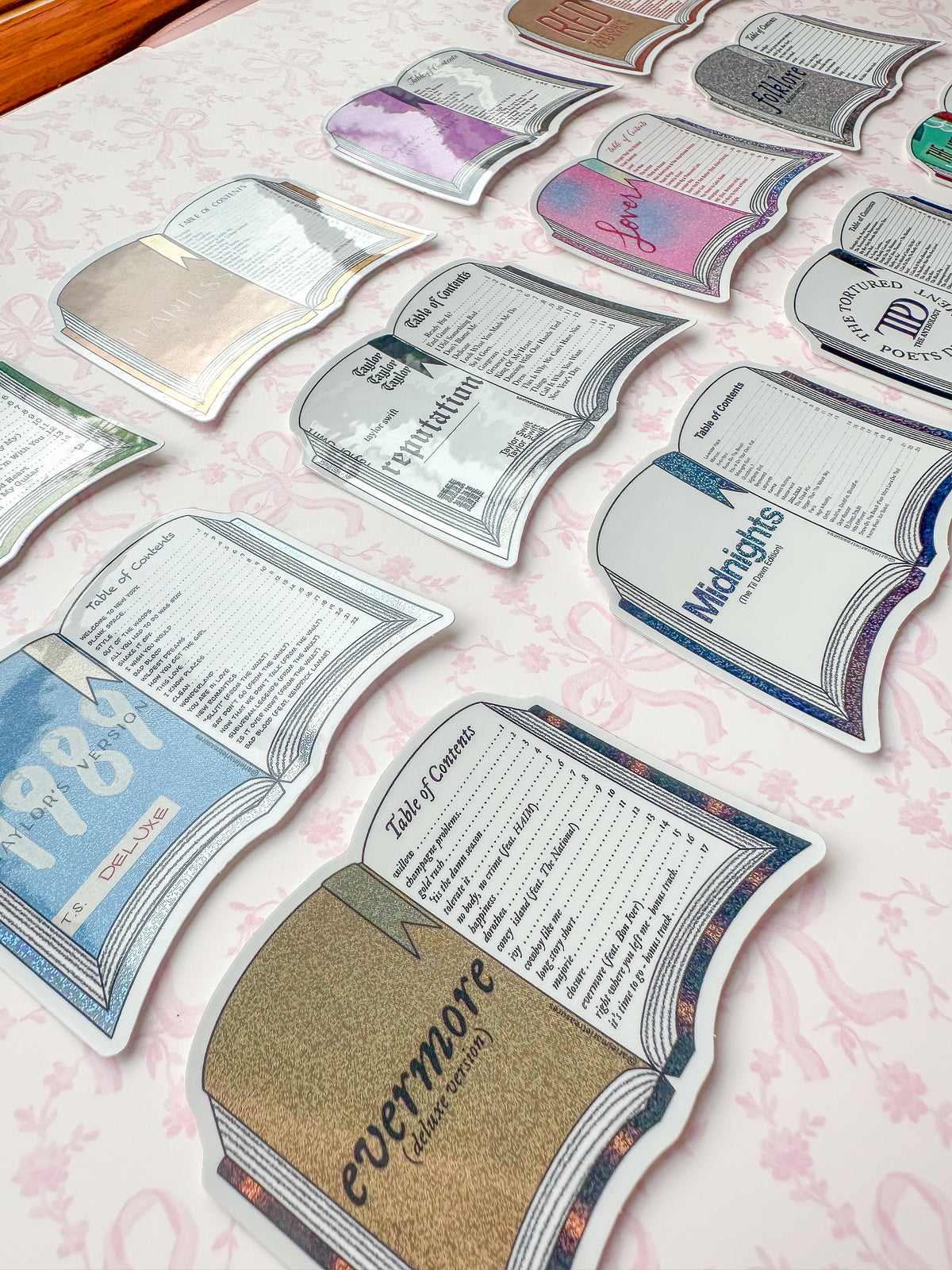 A collection of die-cut book-shaped stickers or cards arranged on a light pink patterned surface. Each book design features different colors and printed text, including titles like '1987', 'Evermore', 'Midnights', and 'the tortured poets department.