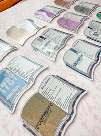 A collection of die-cut book-shaped stickers or cards arranged on a light pink patterned surface. Each book design features different colors and printed text, including titles like '1987', 'Evermore', 'Midnights', and 'the tortured poets department.