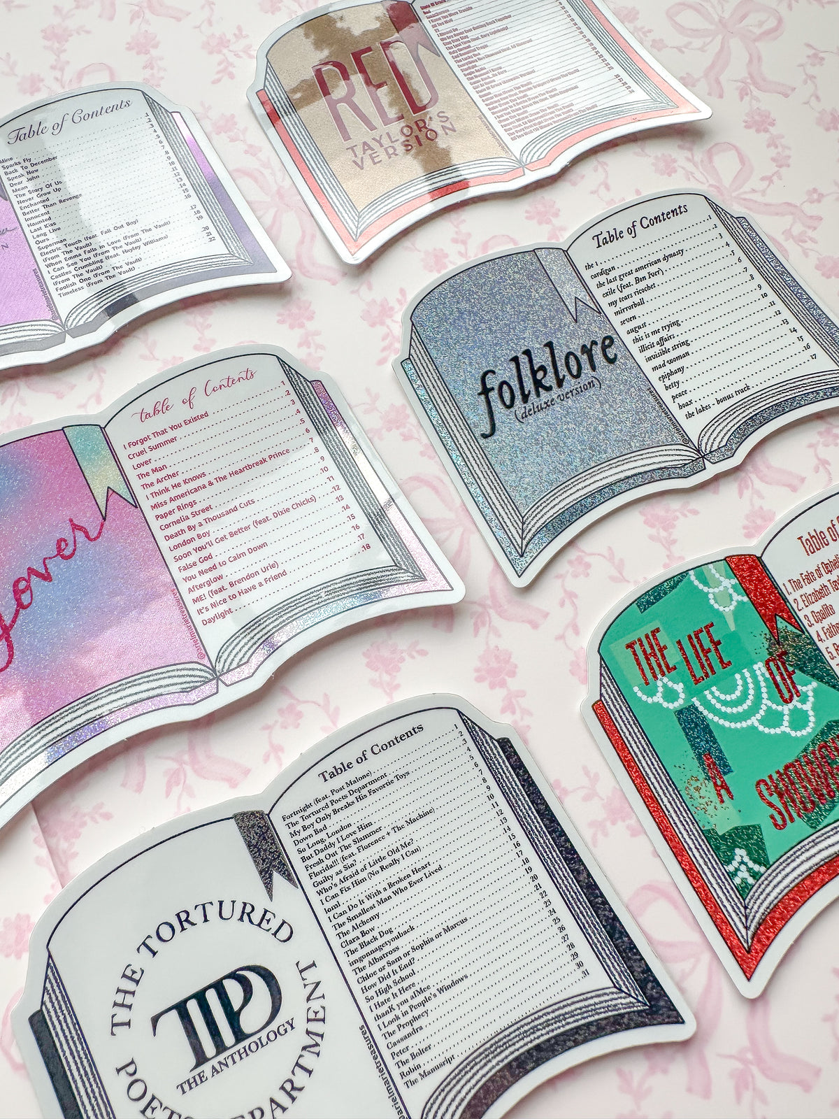A set of die-cut book-shaped table toppers or signs arranged on a pale pink patterned surface. The designs include illustrated open books with titles such as 'Red ' 'folklore ,' 'The Tortured Poet Anthology,' 'The Life of showgirl and 'lover,' each with printed text sections inside.