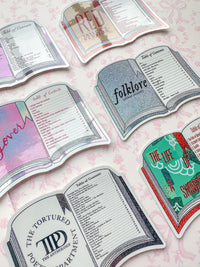 A set of die-cut book-shaped table toppers or signs arranged on a pale pink patterned surface. The designs include illustrated open books with titles such as 'Red ' 'folklore ,' 'The Tortured Poet Anthology,' 'The Life of showgirl and 'lover,' each with printed text sections inside.