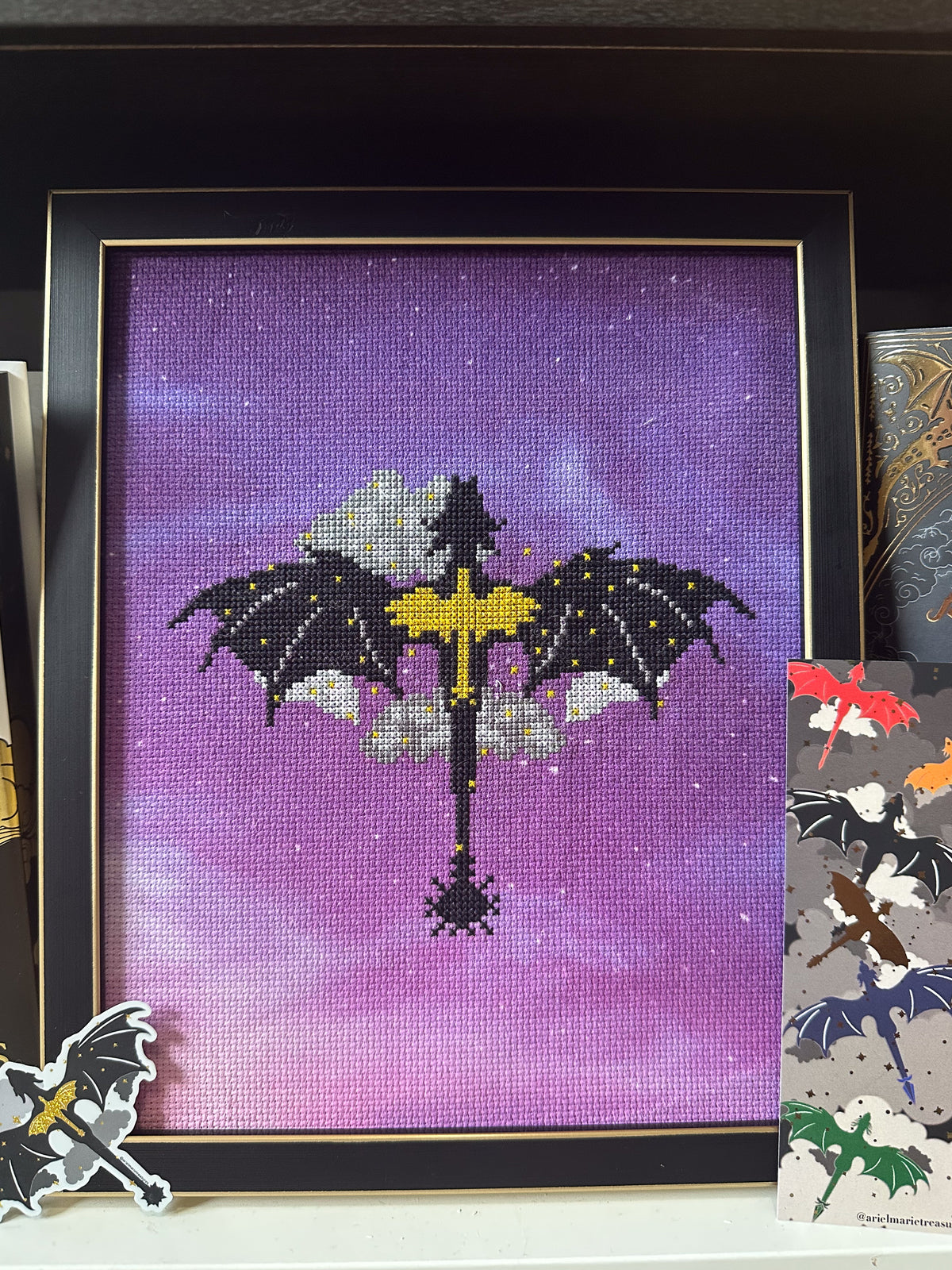 Fourth Wing-inspired cross stitch featuring black dragons, silver clouds, and a golden sword on purple fabric, displayed in a black and gold frame with coordinating dragon stickers.