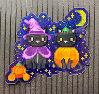 A die-cut sticker featuring a pixelated art design of two black cats dressed in purple Halloween costumes, one wearing a witch hat and the other holding a pumpkin, set against a purple starry night background with a yellow crescent moon.