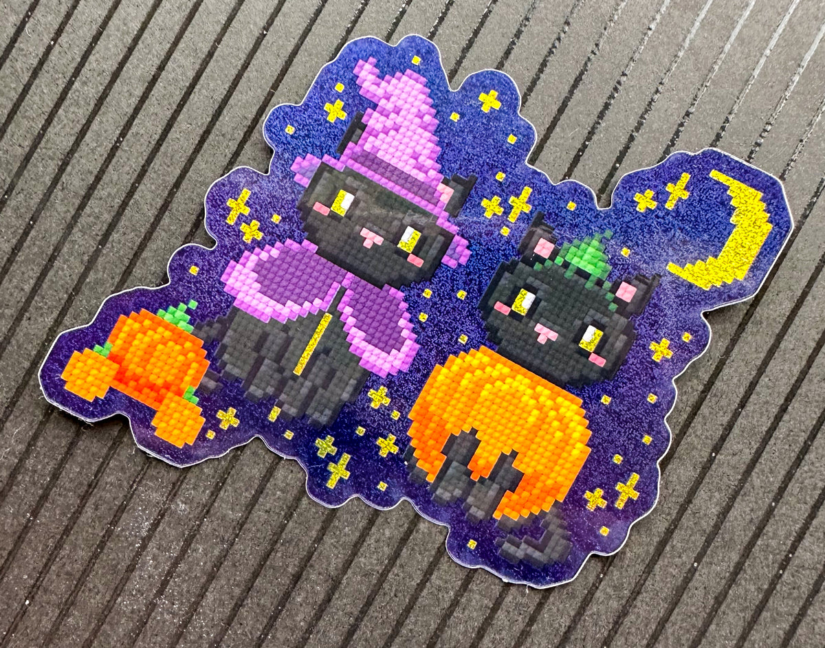 A die-cut sticker featuring a pixelated Halloween design with two black cats, one wearing a pink witch hat and purple dress, and the other holding an orange pumpkin, set against a purple starry night sky. The sticker is placed on a gray ribbed surface.