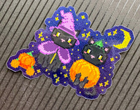 A die-cut sticker featuring a pixelated Halloween design with two black cats, one wearing a pink witch hat and purple dress, and the other holding an orange pumpkin, set against a purple starry night sky. The sticker is placed on a gray ribbed surface.
