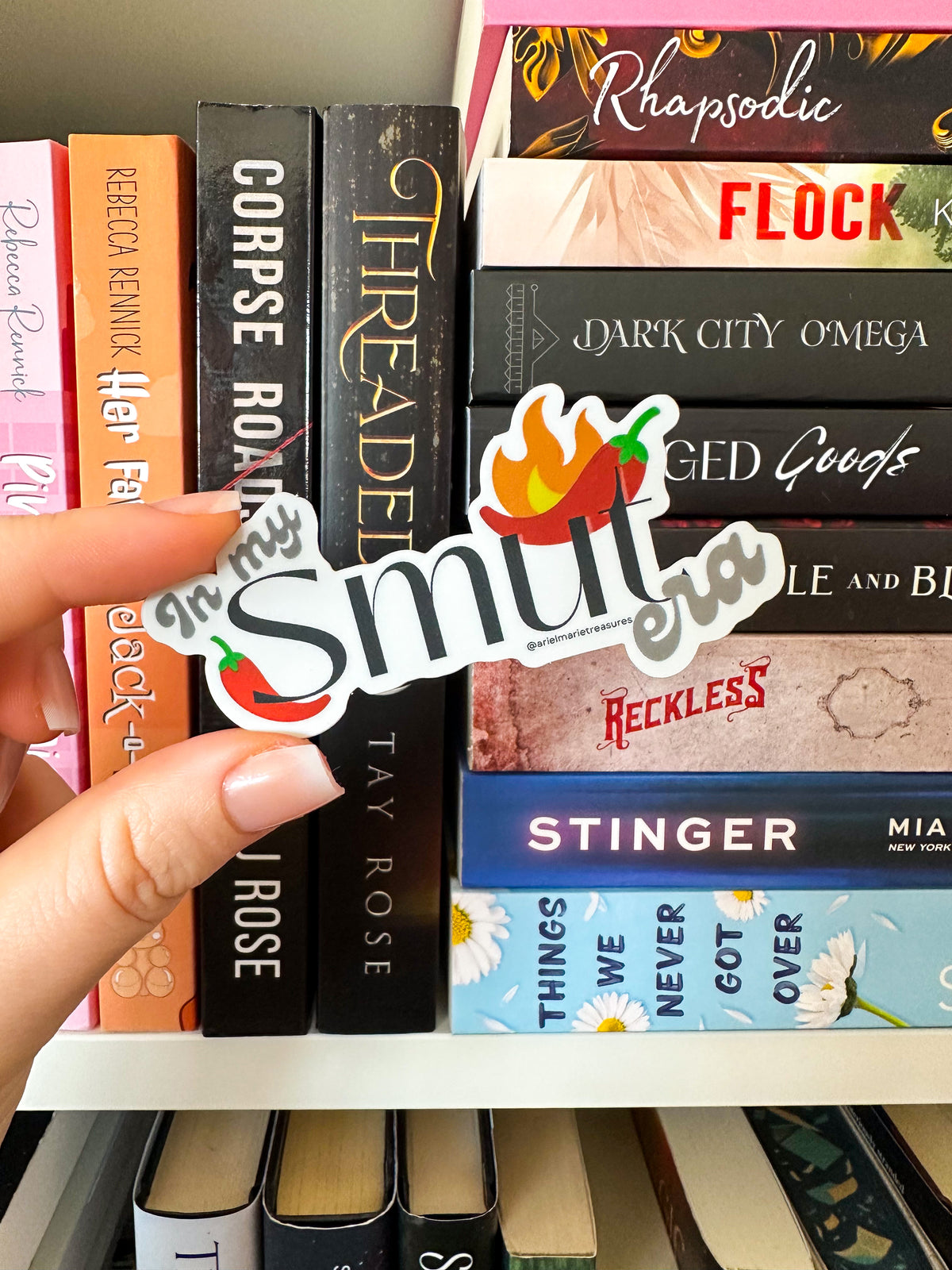 A hand holds a die-cut sticker that reads 'In My Smut' with a chili pepper and flame graphic, positioned in front of a bookshelf filled with romance novels.