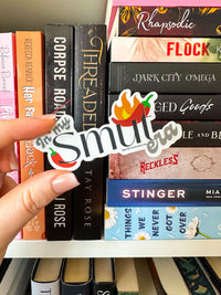 A hand holds a die-cut sticker that reads 'In My Smut' with a chili pepper and flame graphic, positioned in front of a bookshelf filled with romance novels.