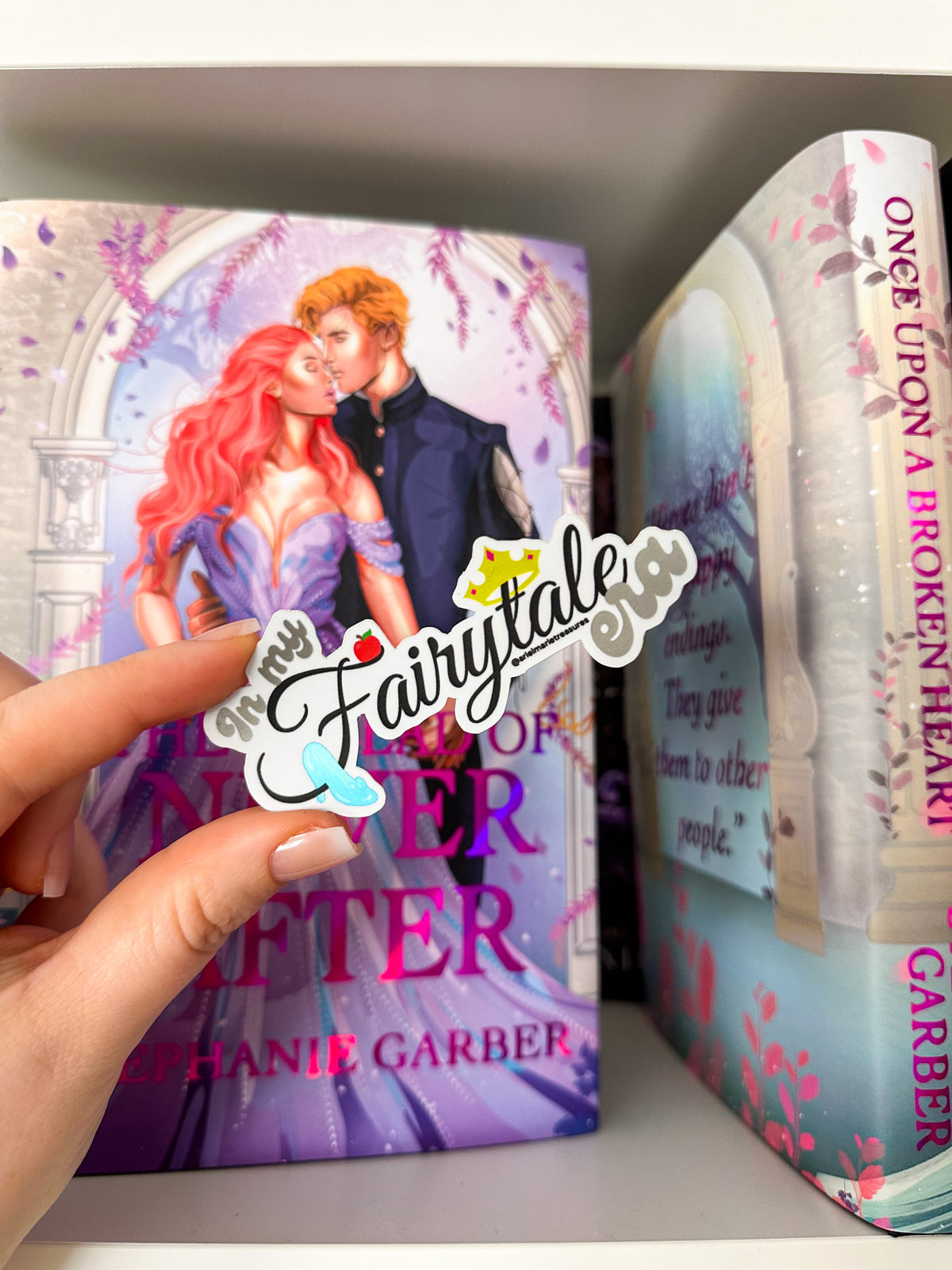 A hand holds a die-cut sticker that reads 'in my Fairytale' in front of a bookshelf with two fantasy books, including 'The Princess of Merlin After' by Stephanie Garber.