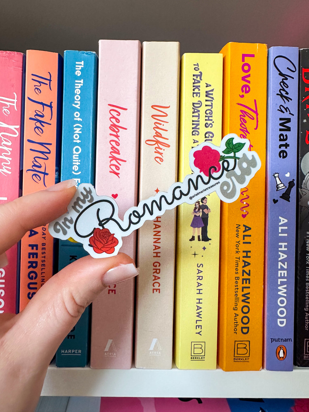 A hand holds a die-cut sticker that reads 'In My Romance Era' in white script with a red rose and green leaves, positioned in front of a row of colorful hardcover books.